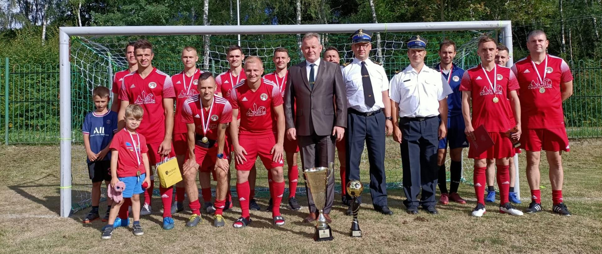 Piłkarze i strażacy stoją w szeregu, w tle bramka piłkarska