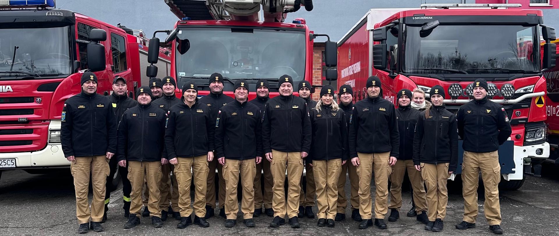 Na zdjęciu znajdują się strażacy ustawieni do zdjęcia na tle samochodów strażackich