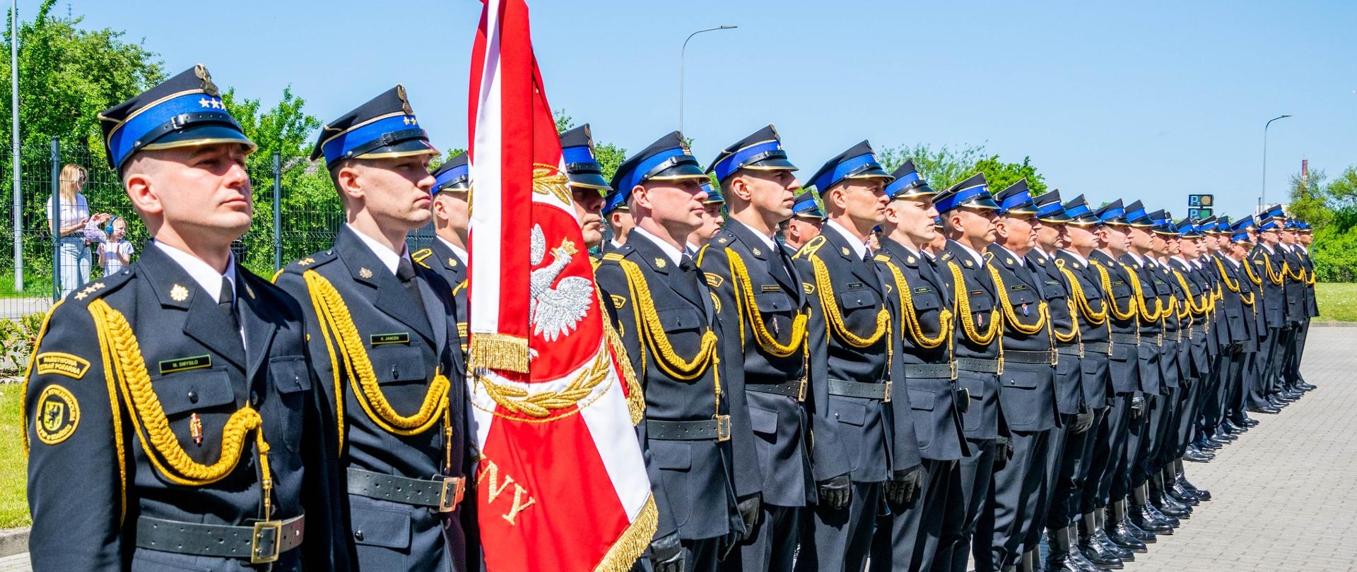 „Zdjęcie przedstawia kompanię honorową Państwowej Straży Pożarnej w Słupsku podczas uroczystych obchodów Dnia Strażaka. Strażacy w galowych mundurach stoją w szyku na placu miejskim, oddając hołd tradycjom pożarniczym i służbie na rzecz bezpieczeństwa mieszkańców.”