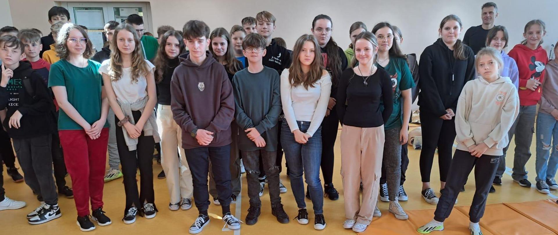 Zdjęcie grupowe , uczestnicy spotkania stoją wraz ze strażakiem na podłodze koloru żółtego. 