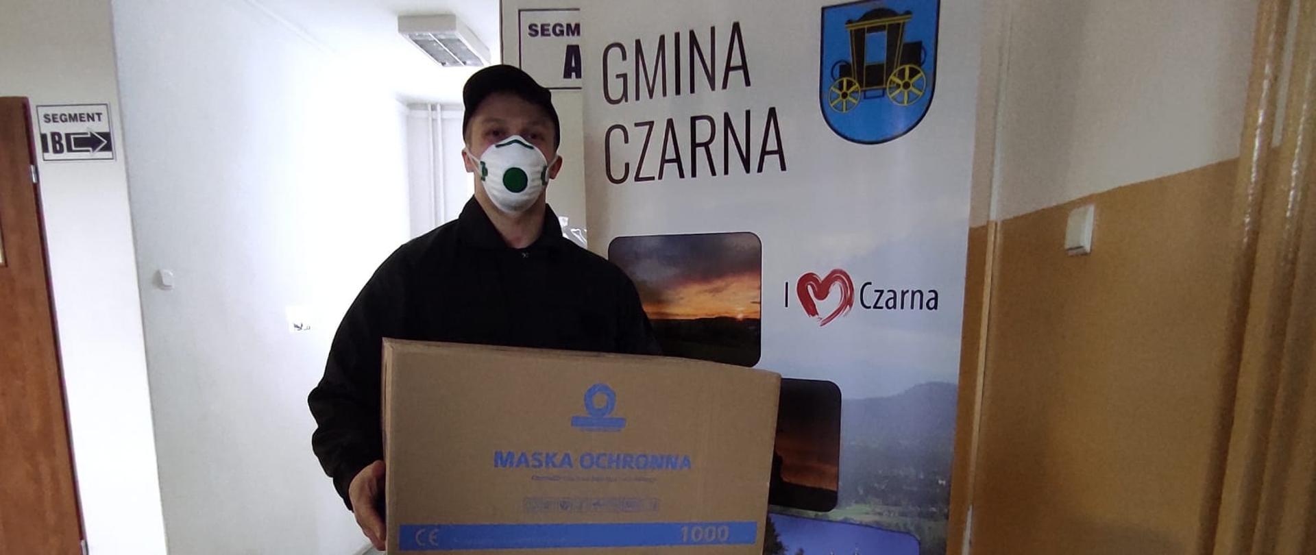 Na zdjęciu strażak PSP w umundurowaniu koszarowym trzymający pudło z maseczkami. W tle baner gminy czarna