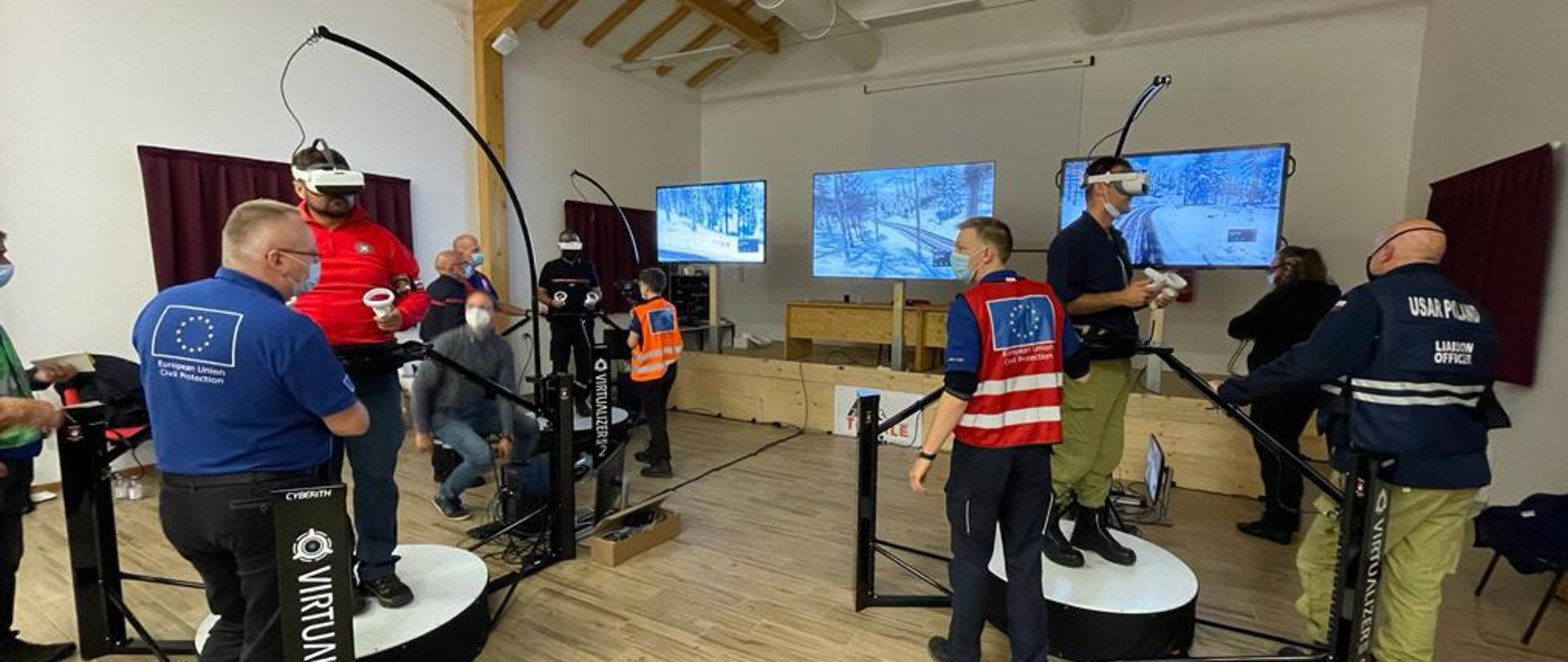 W sali ćwiczeń uczestnicy, dwóch stoi na podestach w okularach VR, na ścianie wiszą 3 ekrany z widokiem z miejscowości gdzie zeszła lawina
