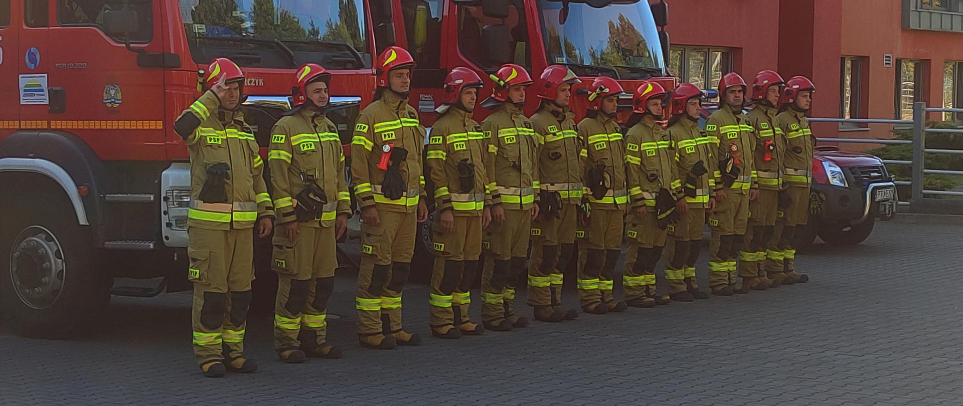 Strażacy ustawieni przed samochodami pożarniczymi.