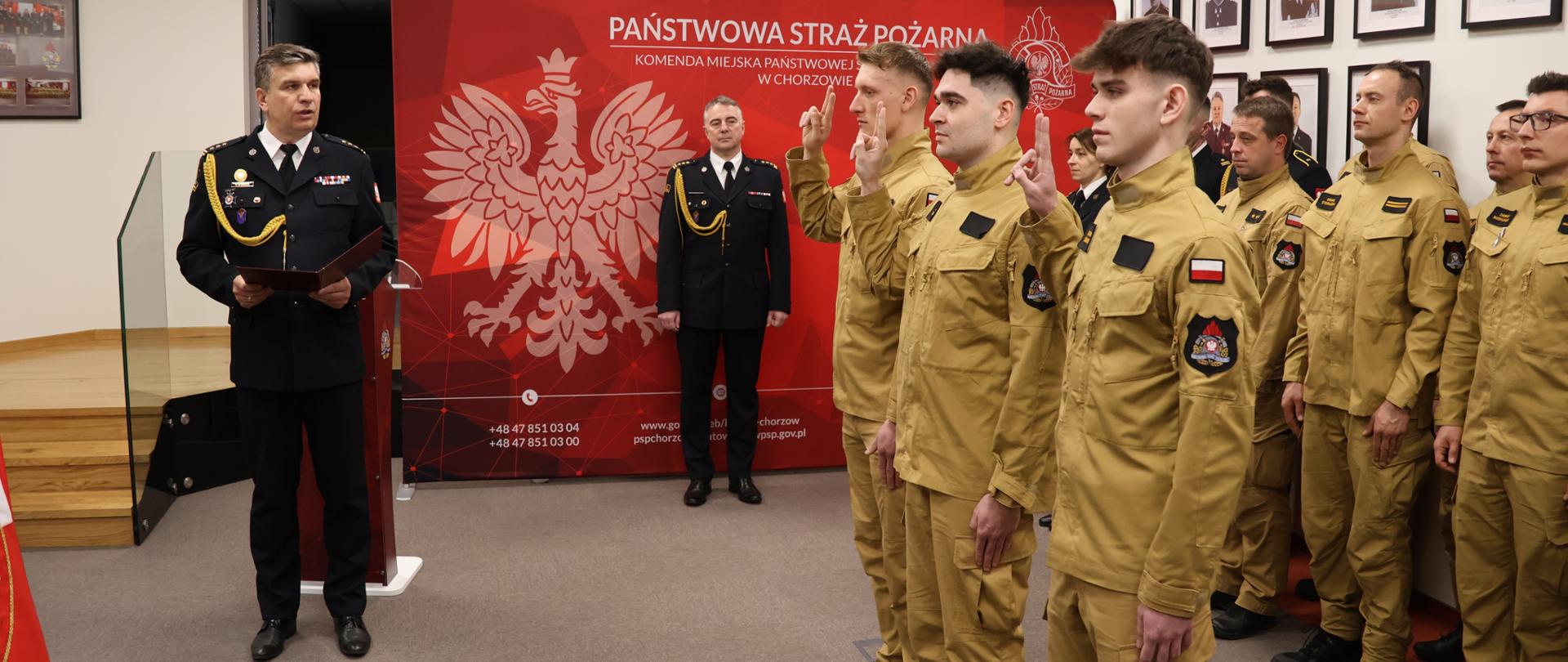 Grupa strażaków Państwowej Straży Pożarnej w galowych mundurach stoi w sali podczas uroczystego ślubowania. Kilku funkcjonariuszy unosi prawe ręce, składając przysięgę. Na pierwszym planie dwóch oficerów w czarnych mundurach prowadzi ceremonię, jeden z nich czyta dokument. W tle widoczna jest czerwona ściana z godłem Polski oraz napisem „Komenda Miejska Państwowej Straży Pożarnej w Chorzowie”.