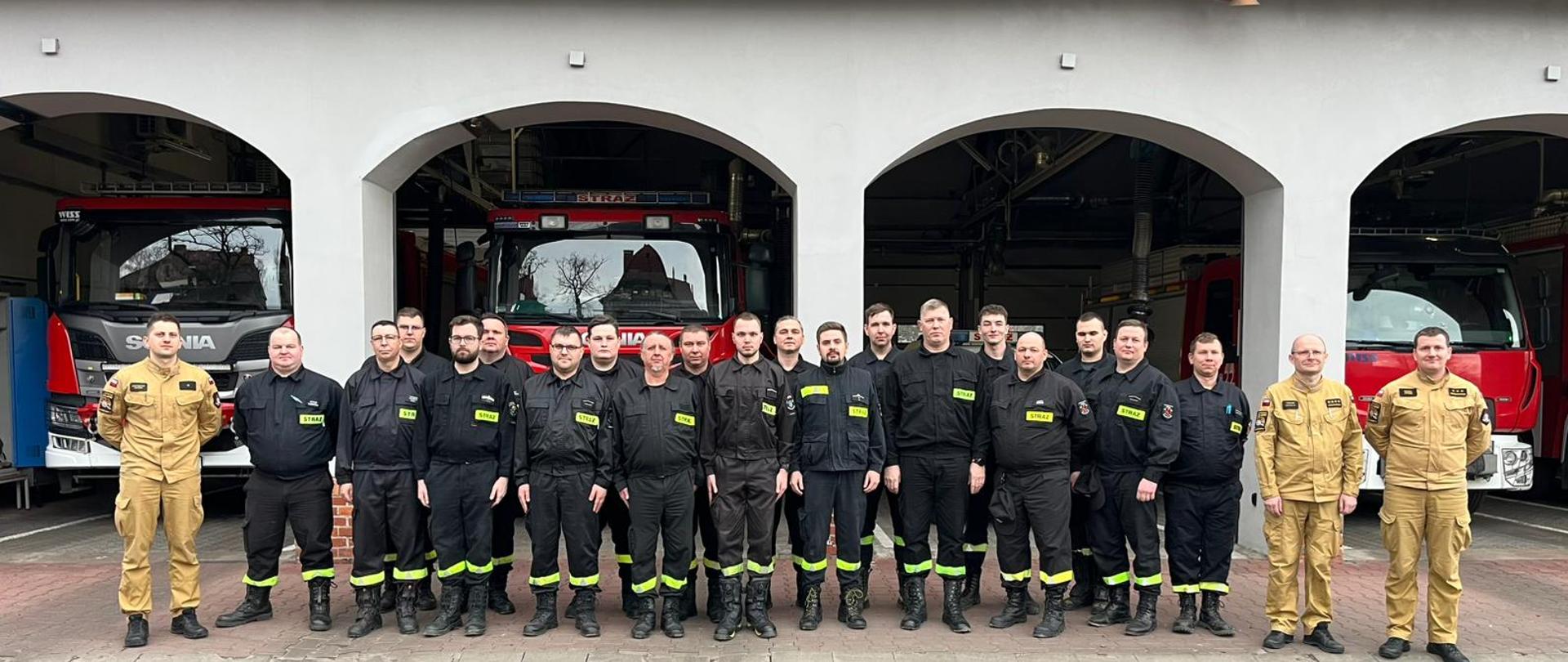 Przed remizą strażacką stoi grupa kilkunastu strażaków w czarnych i beżowych mundurach z odblaskami i naszywkami ‘STRAŻ’. Za nimi, w otwartych bramach garażowych, widoczne są czerwone wozy strażackie marki Scania.