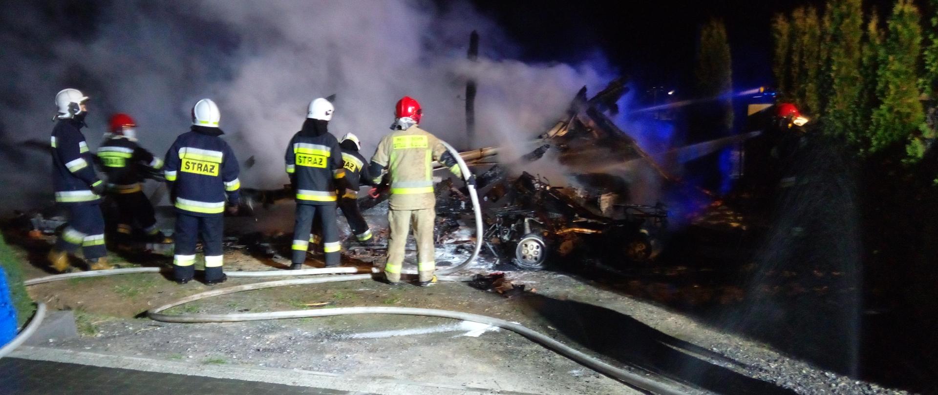 Na zdjęciu widać sześciu strażaków, którzy podając dwa prądy wody dogaszają palącą się wiatę garażową. W oddali widać unoszące się kłęby dymu