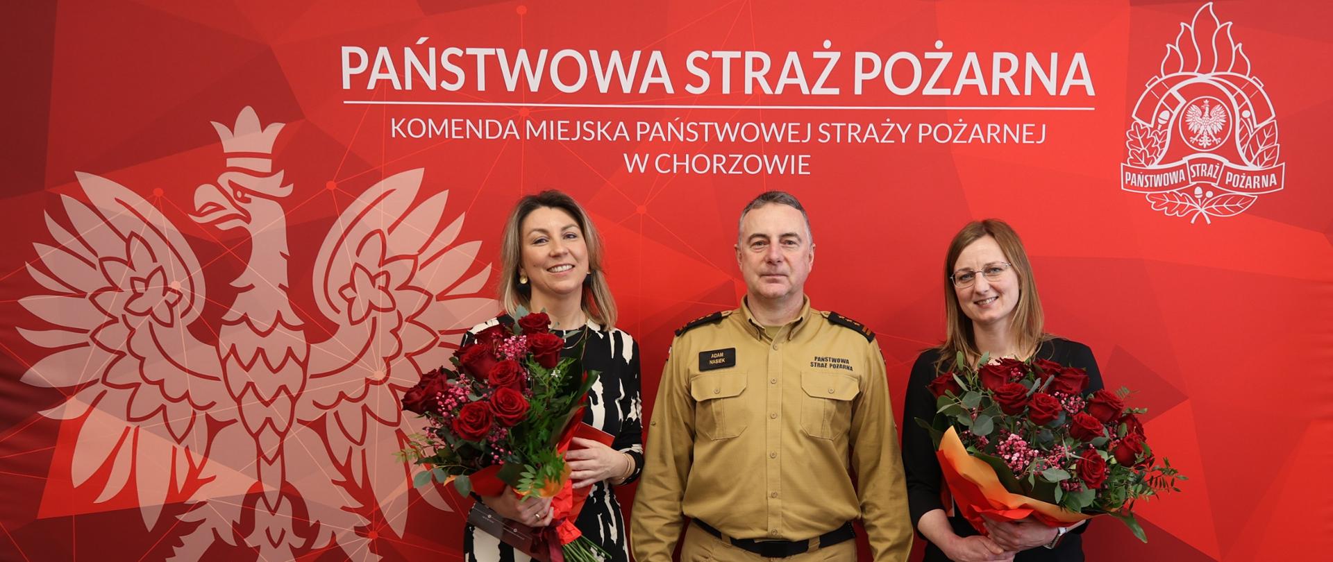 Trzy osoby stoją na tle czerwonej ścianki z emblematem Państwowej Straży Pożarnej oraz nazwą Komendy Miejskiej PSP w Chorzowie. Pośrodku znajduje się mężczyzna Zastępca Komendanta w beżowym mundurze strażackim, a po jego bokach dwie kobiety w eleganckich strojach, trzymające duże bukiety czerwonych róż, przedstawiciele korpusu służby cywilnej. Scena ma charakter uroczysty i nawiązuje do obchodów Święta Korpusu Służby Cywilnej.