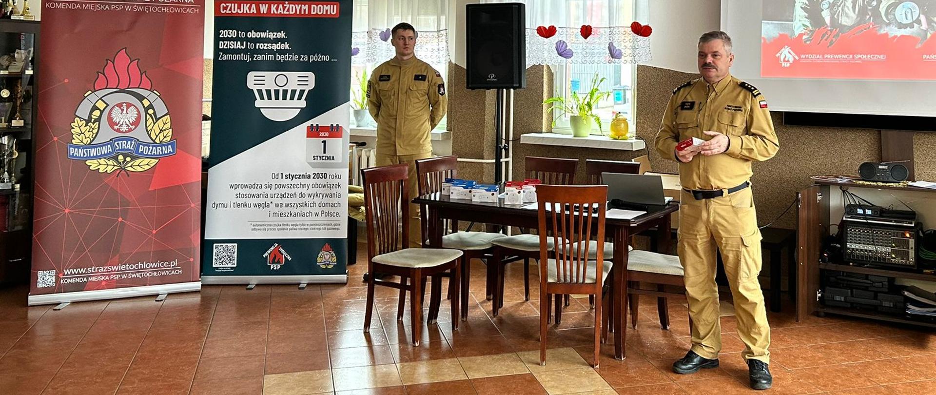 Zdjęcie przedstawia spotkanie edukacyjne zorganizowane przez Państwową Straż Pożarną, w Świętochłowicach. W centrum uwagi znajduje się strażak w piaskowym mundurze, który prowadzi prezentację przed grupą osób, trzymając w dłoniach małe, czerwone opakowanie. W tle widoczny jest ekran z hasłem „Czujka na straży Twojego bezpieczeństwa” oraz banery informujące o obowiązku montażu czujek dymu i tlenku węgla.
Pomieszczenie ma charakter świetlicy z terakotową podłogą i dużymi oknami z firankami. Na stołach rozłożone są materiały edukacyjne i pudełka z urządzeniami, co sugeruje praktyczny pokaz bezpieczeństwa pożarowego. Atmosfera wydaje się formalna, ale przystępna, skupiona na profilaktyce i ochronie życia mieszkańców.
