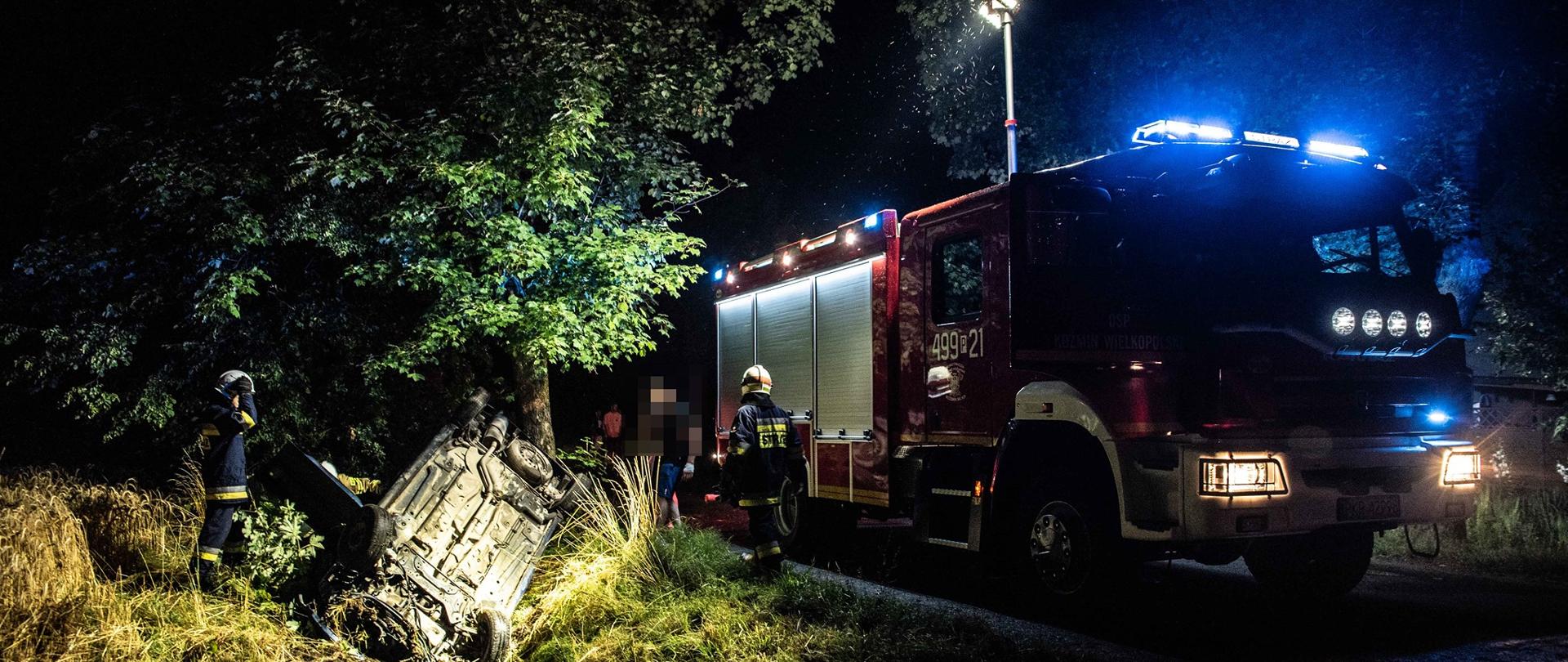 Zdjęcie przedstawia samochód osobowy w rowie w pozycji na dachu w tle samochód pożarniczy i strażacy podczas działań