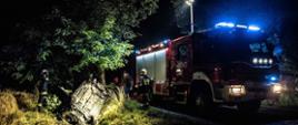 Zdjęcie przedstawia samochód osobowy w rowie w pozycji na dachu w tle samochód pożarniczy i strażacy podczas działań