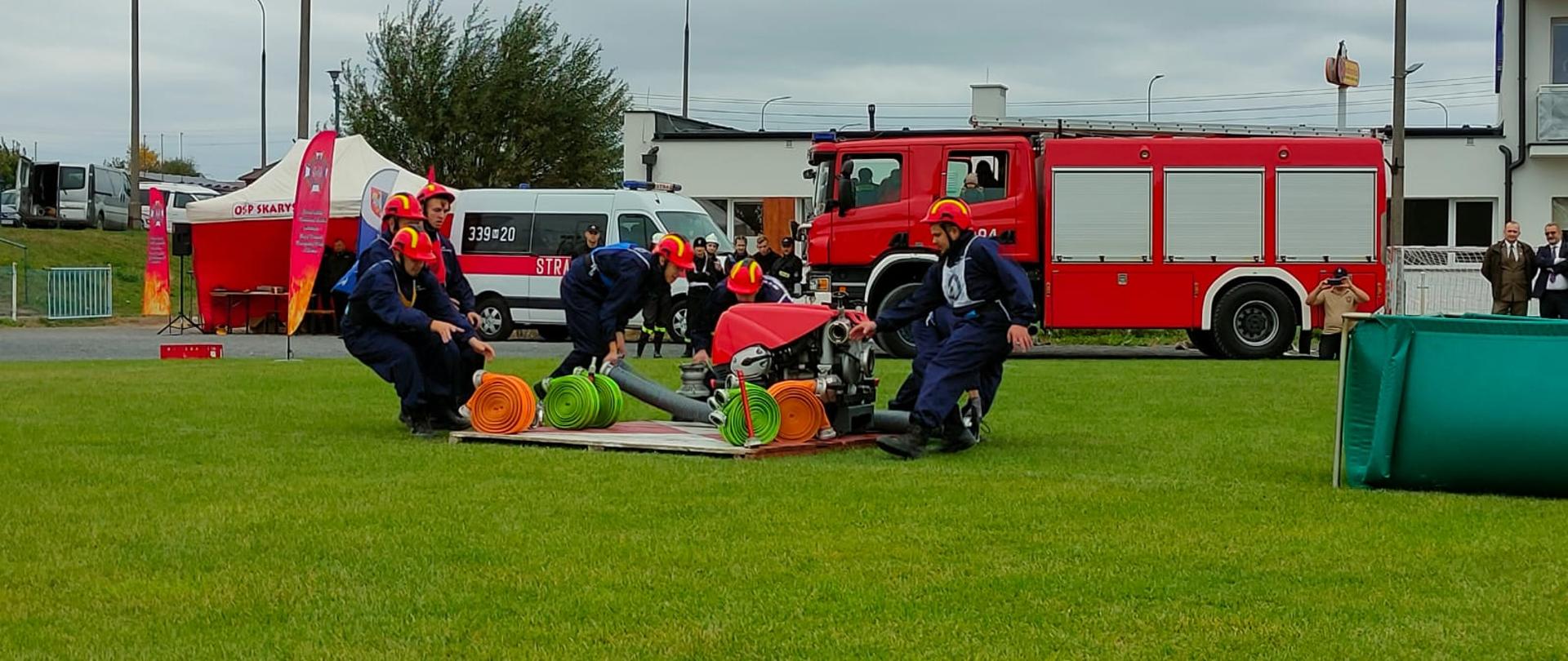 Zdjęcie wykonane podczas rozgrywania zawodów sportowo – pożarniczych jednostek ochotniczych straży pożarnych z terenu powiatu radomskiego. Zawody zostały rozegrane na terenie boiska piłkarskiego w miejscowości wierzbica. Na zdjęciu widać rozpoczynjąca konkurencję rozwinięcie bojowe drużynę. Zawodnicy ubrani są w stroje w kolorze niebieskim. Na głowach założone mają hełmy w kolorze czerwonym. Pomiędzy zawodnikami znajduje się drewniany podest na którym stoi motopompa pożarnicza oraz znajdują się kolorowe węże pożarnicze i inny sprzęt wykorzystywany w tej konkurencji. Po prawej stronie zdjęcia widać zielony zbiornik wykonany z tworzywa sztucznego napełniony wodą. W oddali za zawodnikami widać pojazdy pożarnicze oraz ustawiony namiot organizatora zawodów. 