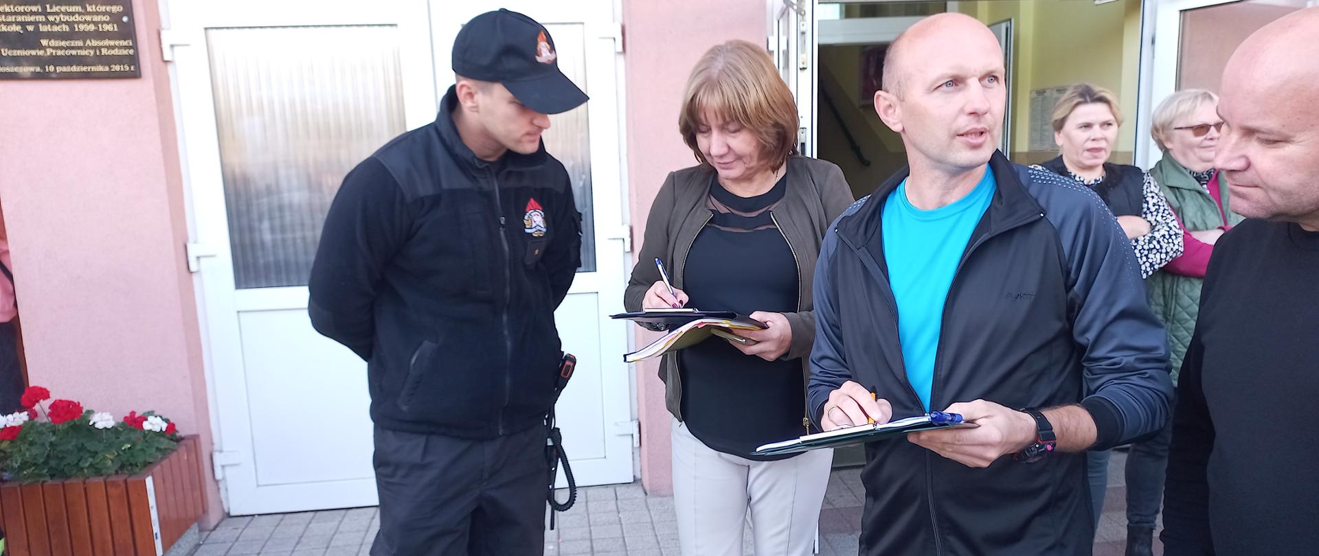 Wejście do budynku szkoły, strażak z pracownikami szkoły podczas przeliczenia młodzieży w trakcie próbnej ewakuacji.
