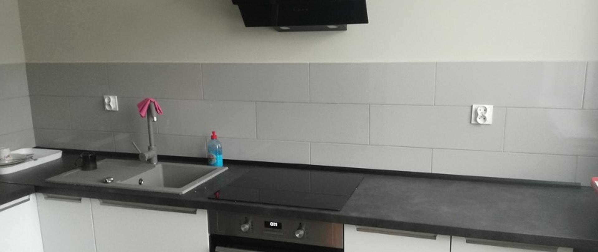 Zdjęcie przedstawia kuchenne pomieszczenie socjalne. Widok białych mebli kuchennych ustawionych na terakocie. W centralnym punkcie zdjęcia czarny okap kuchenny nad kuchnią indukcyjną. Z lewej strony czarnej płyty indukcyjnej zlew jednokomorowy z baterią. Ściany wypełnione trzema warstwami prostokątnych, szarych płytek