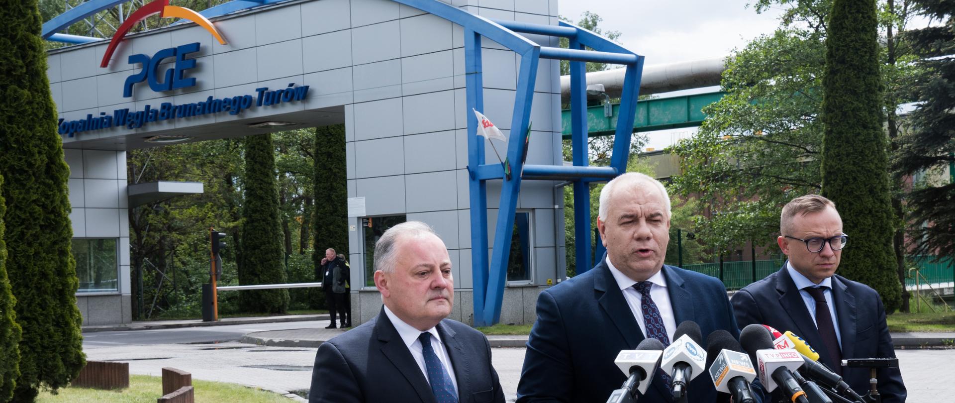 Wicepremier Jacek Sasin w towarzystwie prezesa PGE Wojciecha Dąbrowskiego (po lewej) oraz wiceministra Artura Sobonia (po prawej) W tle brama wjazdowa do kopalni, z napisem: Kopalnia Węgla Brunatnego Turów i logotypem spółki.