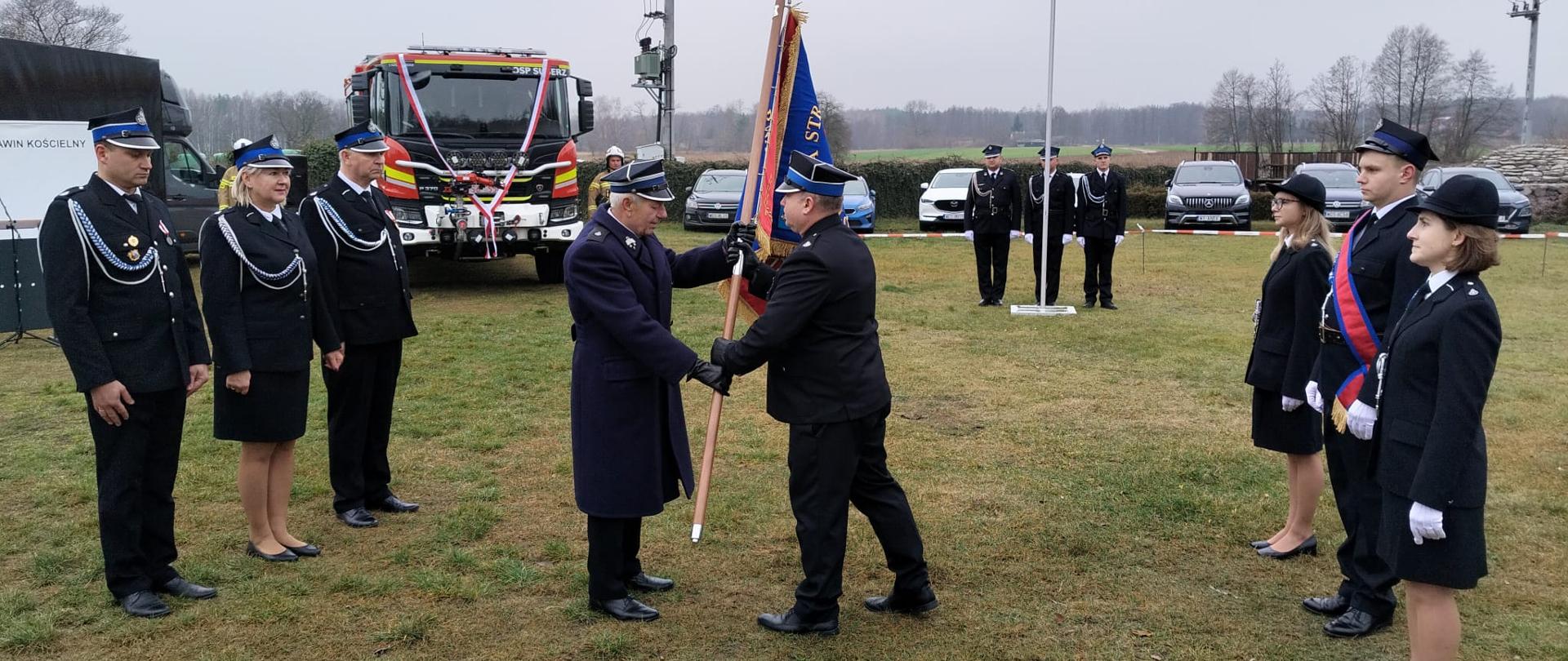 Na zdjęciu strażacy OSP przekazują sobie sztandar. W tle czerwony samochód strażacki i kilka samochodów osobowych. Strażacy stoją na trawie, powyżej widoczne jest pochmurne niebo.