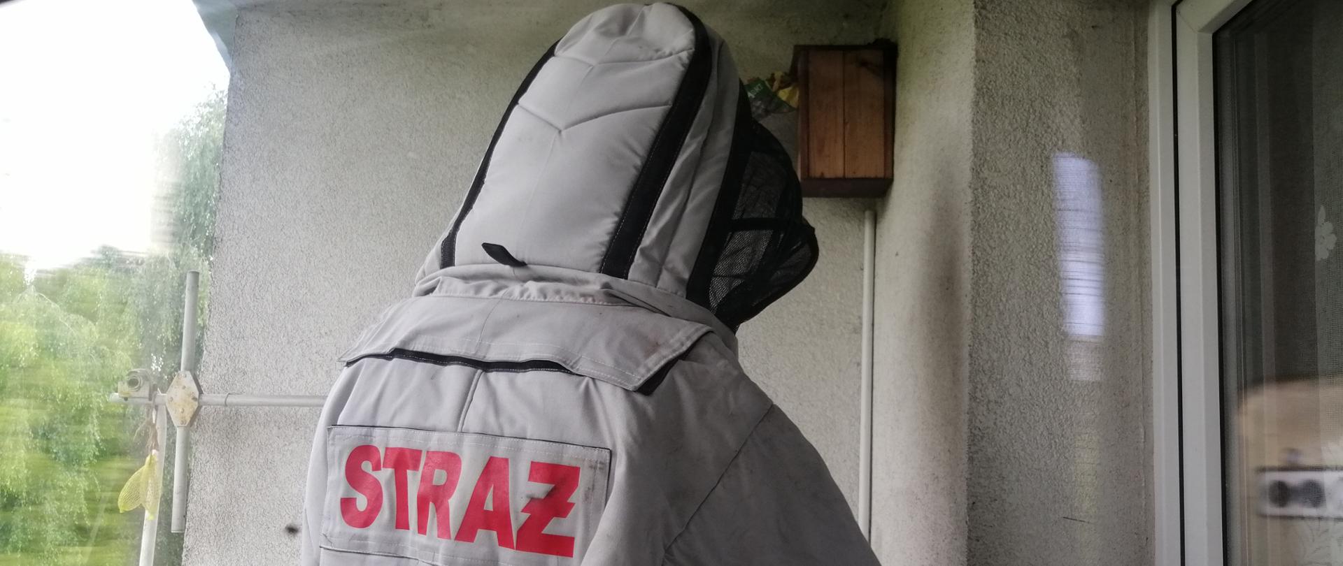 Zdjęcie przedstawia osobę ubrana w szary kombinezon z czerwonym napisem STRAŻ na plecach, oraz z czarną siatką na twarz. Osoba ta stoi na balkonie jest zwrócona plecami do fotografa. 