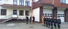 Na zdjęciu widoczni strażacy na tle budynku komendy. Na środku zdjęcia masz z flagą i salutujący trzej strażacy. Po prawej stronie zmiana służbowa w jednym szeregu z salutującym dowódcą zmiany służbowej.