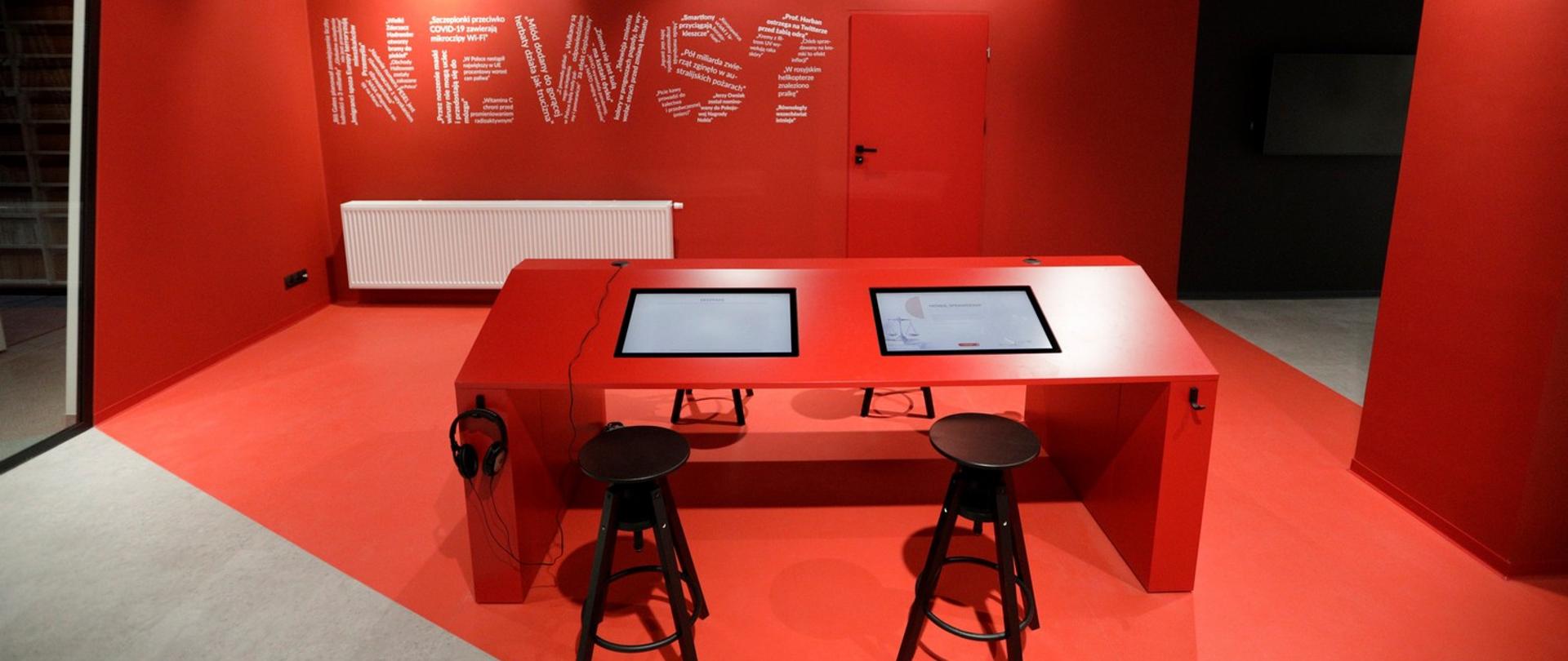 News room w centrum edukacji medialnej. Pokój z czerwonymi ścianami, pośrodku prostokątny stół cztery, wysokie taborety. W blacie stołu wbudowane dwa monitory.