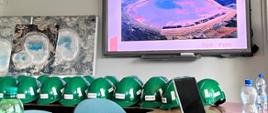 Zdjęcie przedstawia wnętrze sali szkoleniowej z dużym ekranem multimedialnym wyświetlającym prezentację KGHM. Na stole w dolnej części kadru ułożony jest długi rząd zielonych kasków ochronnych z logo KGHM, a obok nich widoczne są butelki z wodą mineralną oraz laptop.