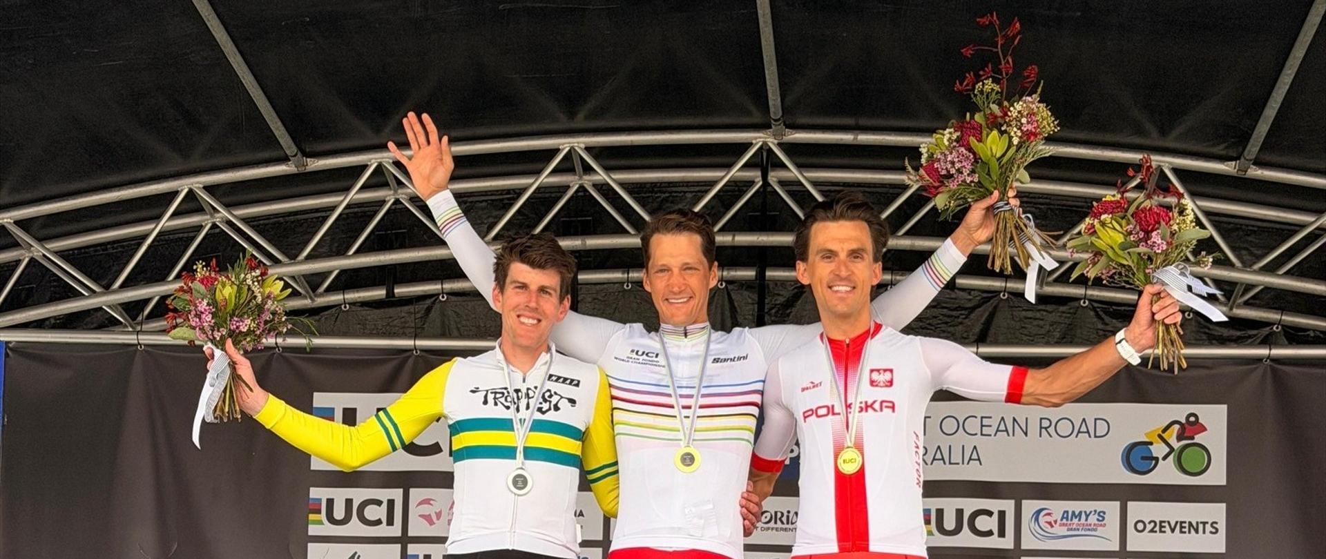 „Trzech kolarzy stoi na podium po wyścigu, każdy trzyma bukiet kwiatów. Na środku zawodnik w białym stroju z medalem złotym, obok niego kolarz w biało-czerwonym stroju reprezentacji Polski z medalem srebrnym.”
