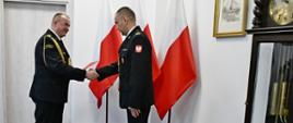 Strażak w mundurze wyjściowym ze sznurem trzyma czerwoną teczkę gratuluje stojącemu przed nim strażakowi w mundurze wyjściowym obok stoją trzy flagi Polski są drzwi na ścianie wisi obraz oraz obok stoi zegar. 