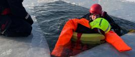 Na zdjęciu ratownik z manekinem w przeręblu, drugi ratownik klęczący przy tafli lodu. 