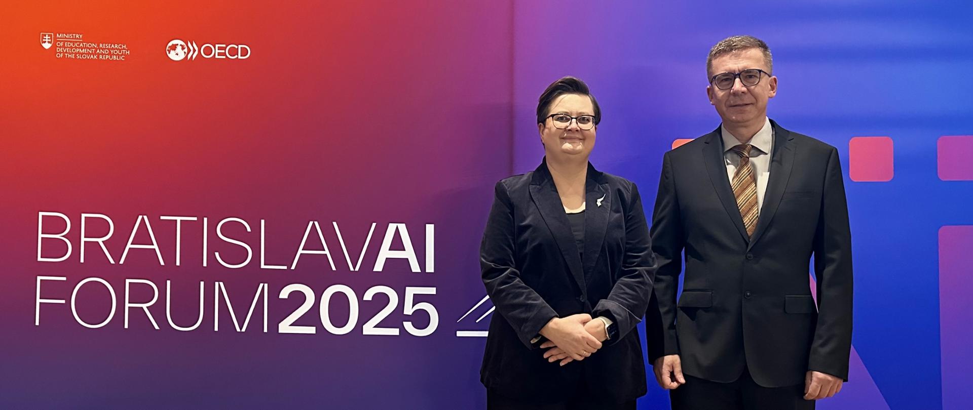 Kobieta w okularach i czarnym garniturze oraz mężczyzna w garniturze i krawacie pozują na tle dużej, niebiesko-fioletowej ściany z napisem „BRATISLAVAI FORUM 2025” oraz logo OECD.
