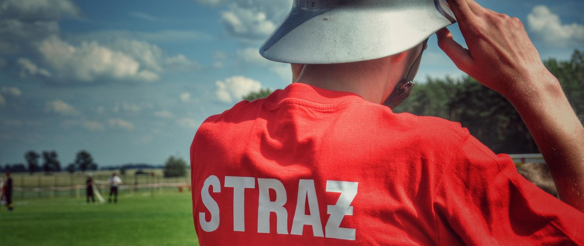 Zdjęcie przedstawiające mężczyznę w jasnym hełmie strażackim. Ma na sobie czerwoną koszulkę z napisem "STRAŻ". Tło przedstawia zielone boisko, a w oddali widać las. Niebo jest częściowo pokryte białymi chmurami.