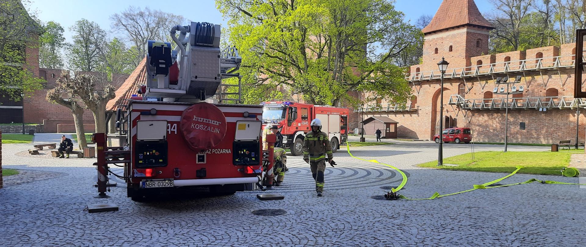 Duży plac pokryty kostką brukową. Podnośnik strażacki przygotowuje się do działań. Strażacy przy samochodzie. Plac otoczony zabytkowymi murami z czerwonej cegły. Słonecznie.