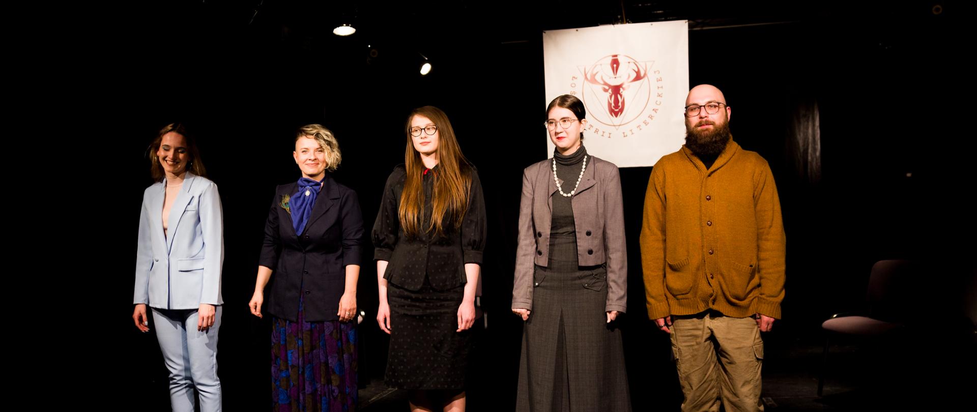 Pięcioro aktorów stojących na czarnej scenie teatralnej po spektaklu. 