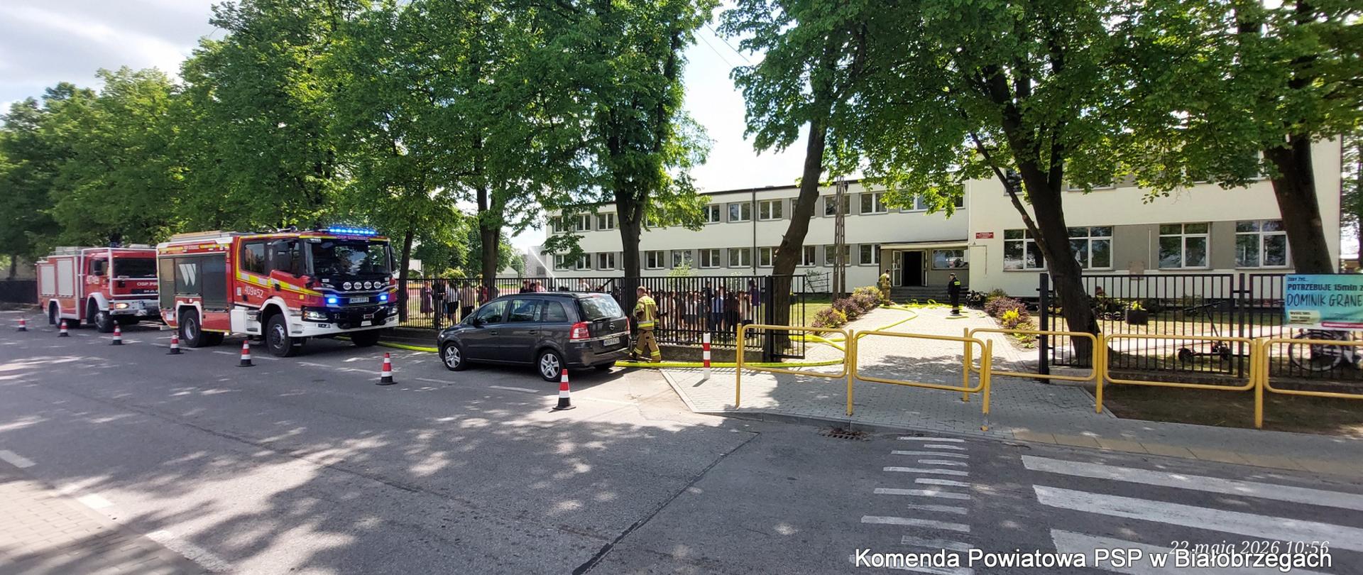 Ujęcie z przeciwnej strony drogi przedstawia widok na budynek Publicznej Szkoły Podstawowej im. Dionizego Czachowskiego w Bobrku w trakcie ćwiczeń. Przed szkolnym ogrodzeniem i żółtą barierą zaparkowane są dwa czerwone wzy do straży pożarnej (jeden z JRG Białobrzegi z napisem "W"). Między nimi stoi grafitowy samochód osobowy. Strażacy w piaskowych mundurach poruszają się po terenie przed szkołą. Droga przed szkołą jest pusta, a na niej widać pasy przejściowe. W prawym dolnym rogu znajduje się biały datownik: "22 maja 2026 10:56".