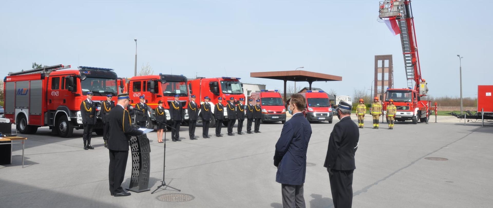 Zdjęcie przedstawia plac wewnętrzny Komendy Powiatowej PSP w Opocznie, na którym trwa uroczystość, strażacy w ubraniach galowych, przemawiający Komendant Powiatowy PSP w Opocznie, zgromadzeni są goście. W tle flaga Rzeczypospolitej Polskiej oraz wystawione pojazdy pożarnicze.