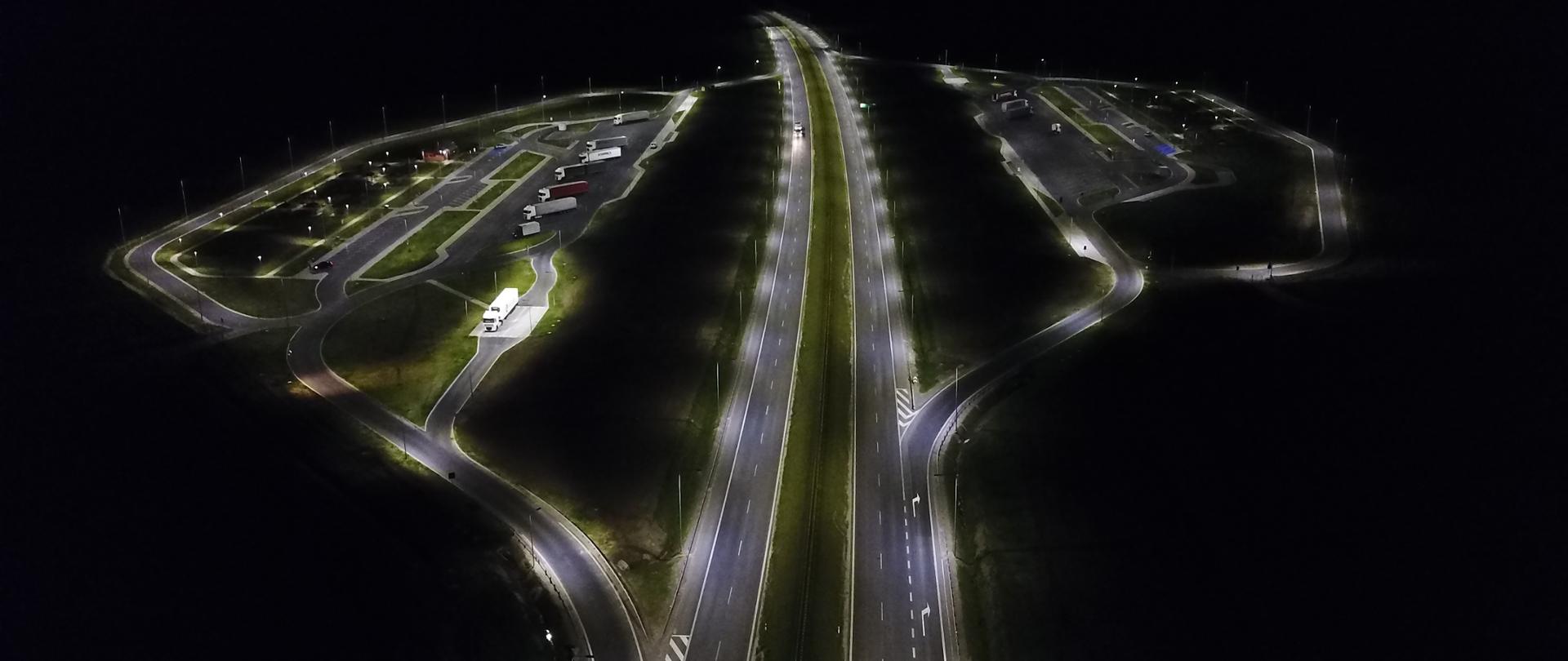 Widok nocny. Po lewej i prawej stromnie widoczne oświetlone miejsca obsługi podróżnych z zaparkowanymi samochodami, po środku widoczna droga ekspresowa dwupasowa.