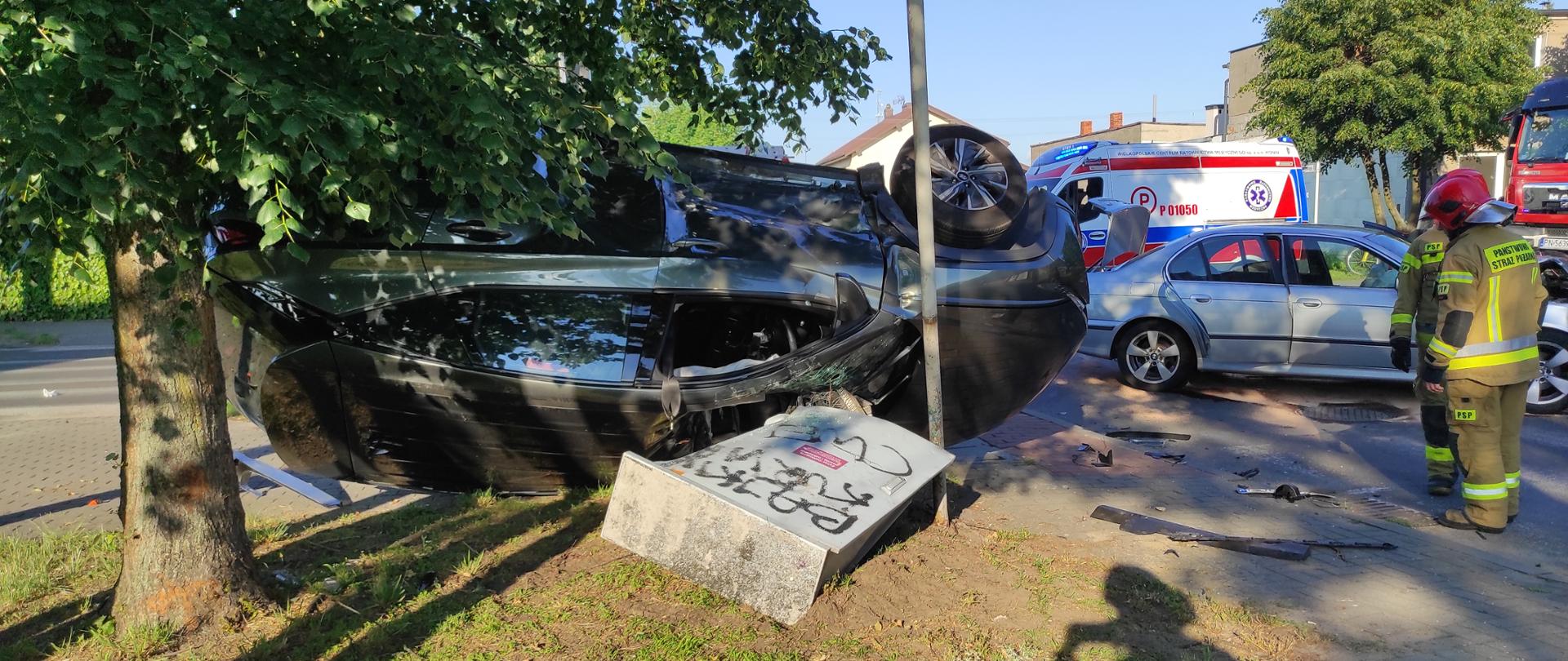 Zdjęcie przedstawia dwa samochody osobowe po wypadku. Jeden z pojazdów leży na dachu opary o skrzynkę energetyczną, drugi stoi na środku skrzyżowania. Ponadto na zdjęciu widać strażaków, karetę pogotowia oraz samochód strażacki. 