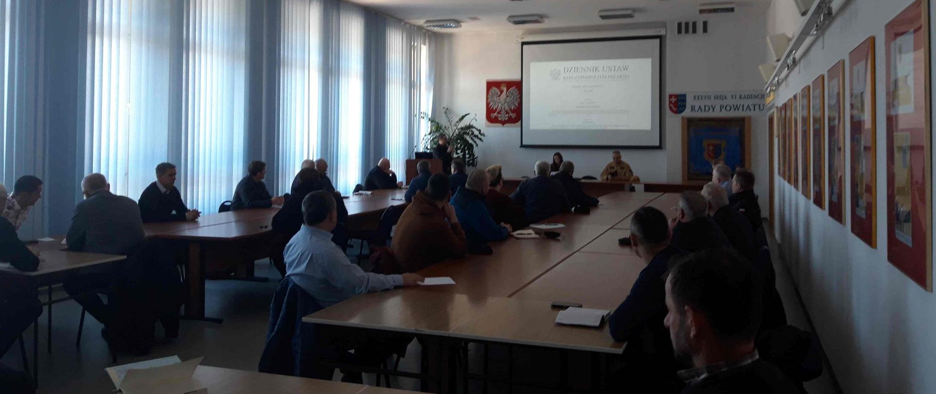 W pomieszczeniu przy stołach siedzi kilkadziesiąt osób. Przed niektórymi na blatach leżą dokumenty. Po lewej stronie ściana z dużymi oknami przesłoniętym roletami, po prawej na ścianie wiele obrazów. Na ścianie na wprost ekran na którym wyświetlany jest tekst – po jego lewej stronie godło Polski, po prawej sztandar.