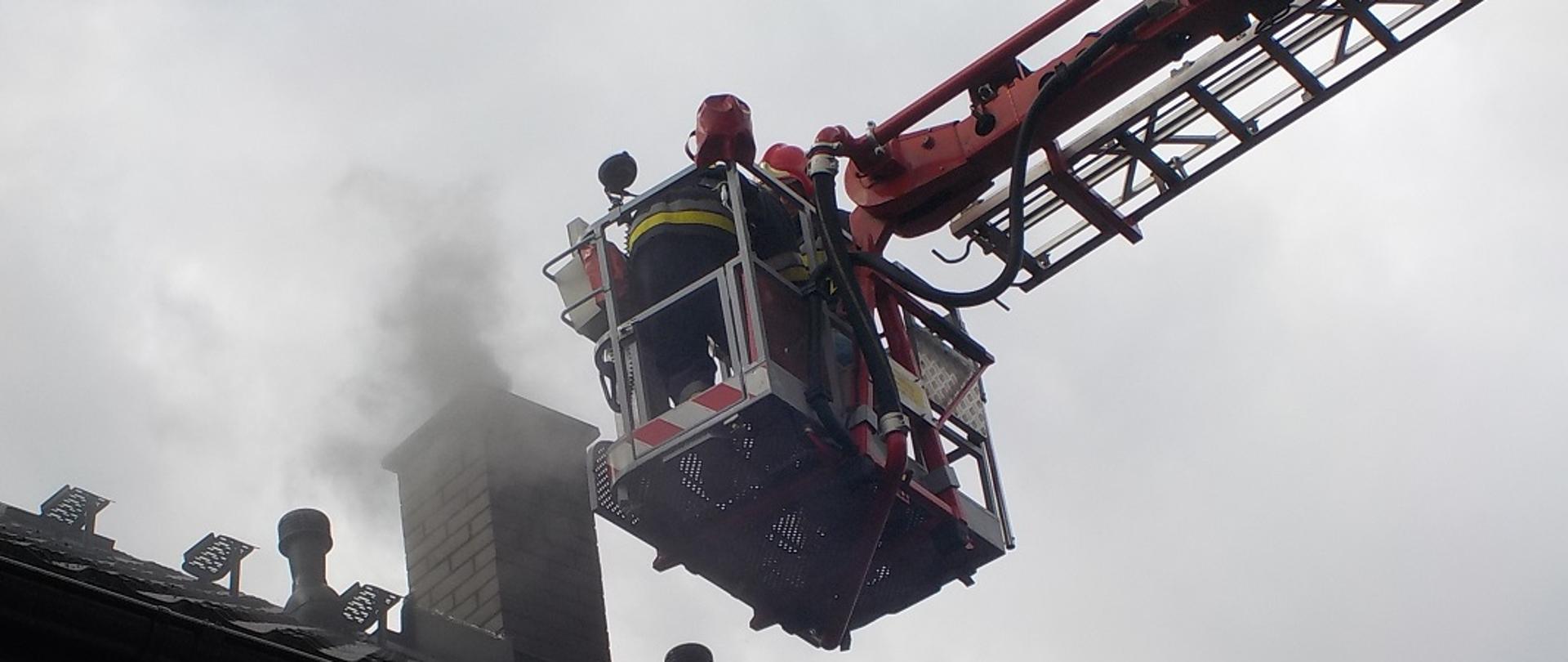 Fotografia przedstawia fragment dachu budynku z widocznym kominem. Przy kominie w koszu podnośnika widać dwóch strażaków w ubraniach bojowych i hełmach podejmujących działania ratowniczo gaśnicze wynikające z zapalenia się sadzy w przewodzie kominowym.