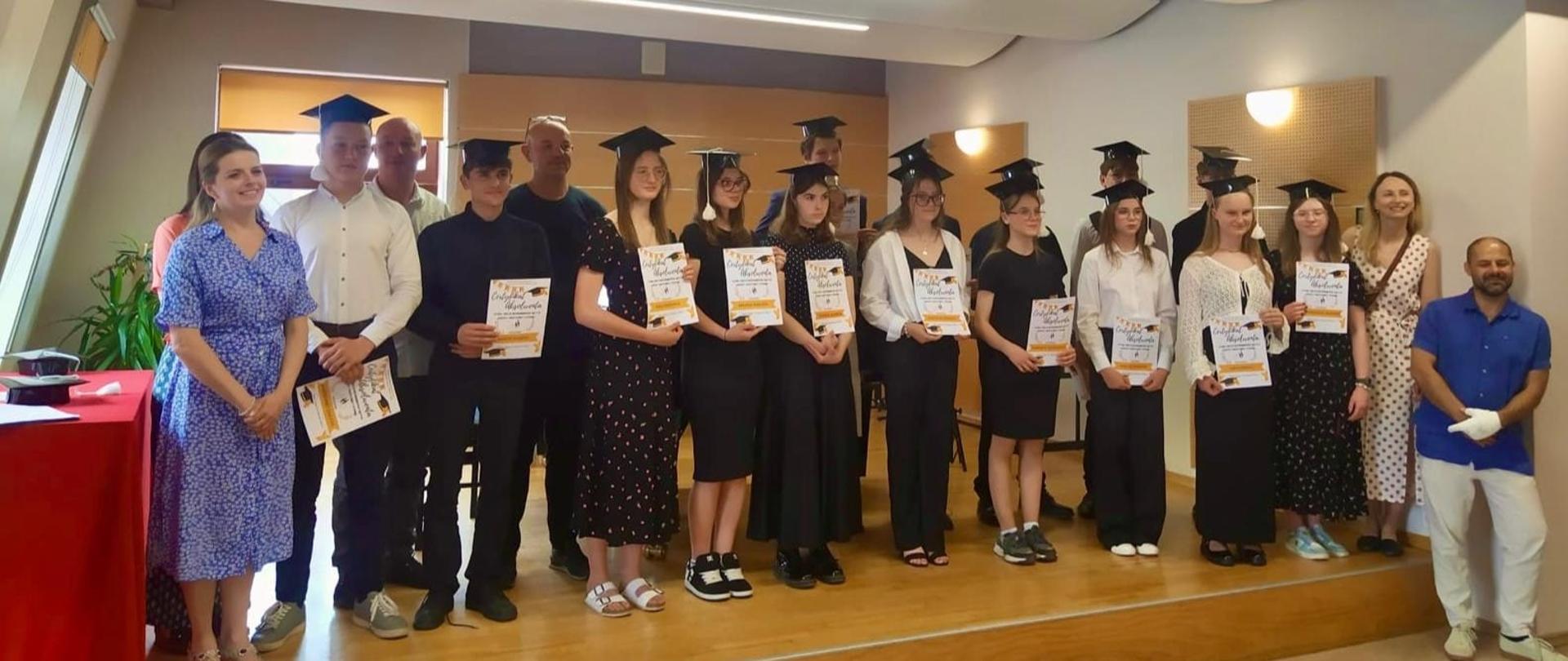 Na zdjęciu widoczna jest uroczystość zakończenie dla uczniów sekcji dętej. Grupa około 20 osób stoi na podwyższeniu – są to głównie młodzież i kilku dorosłych- nauczyciele. Młodzież (około 15 osób, głównie dziewczęta i kilku chłopców) stoi w szeregu na środku sceny. Mają na sobie odświętne stroje – głównie ciemne lub eleganckie ubrania – oraz czarne birety absolwenckie na głowach. W rękach trzymają dyplomy. Po bokach grupy stoją dorośli – czterech nauczycieli lub opiekunów z lewej i trzech z prawej strony. Jedna kobieta ubrana jest w niebieską sukienkę w kwiaty, druga w białą sukienkę w czarne grochy, jeden z mężczyzn ma granatową koszulę i białe spodnie. Po lewej stronie zdjęcia znajduje się czerwony stół, na którym leżą birety i kartki – prawdopodobnie przygotowane do dalszego wręczania. W tle widać jasną, nowoczesną salę z drewnianą podłogą i ścianami, oświetloną światłem dziennym. Atmosfera jest radosna i uroczysta – to moment dumy i zakończenia etapu edukacji.