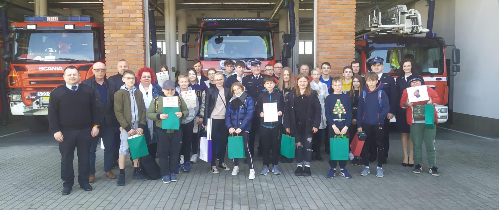 Zdjęcie przedstawia wszystkich uczestników eliminacji powiatowych Ogólnopolskiego Turnieju Wiedzy Pożarniczej „Młodzież Zapobiega Pożarom” z nagrodami/dyplomami oraz opiekunów z poszczególnych szkół i organizatorów przedsięwzięcia, widocznych na tle pojazdów pożarniczych zlokalizowanych w garażach Komendy Powiatowej Państwowej Straży Pożarnej w Gołdapi