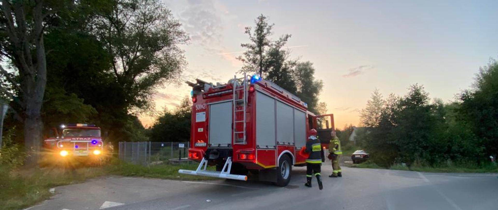 Samochód pożarniczy stoi na łuku drogi przed nim dwóch strażaków.
