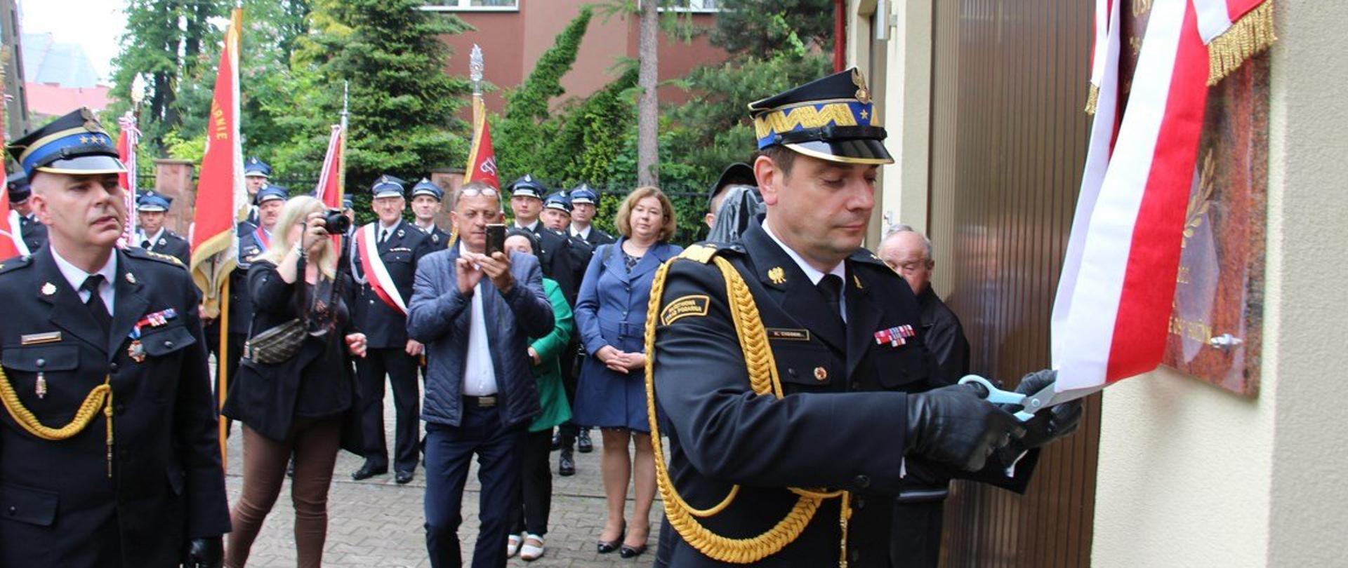 Zdjęcie przedstawia komendanta wojewódzkiego PSP w Kielcach odsłaniającego tablicę pamiątkową z okazji 112-lecia powstania OSP Bliżyn. W tle widać przybyłych gości i sztandary.