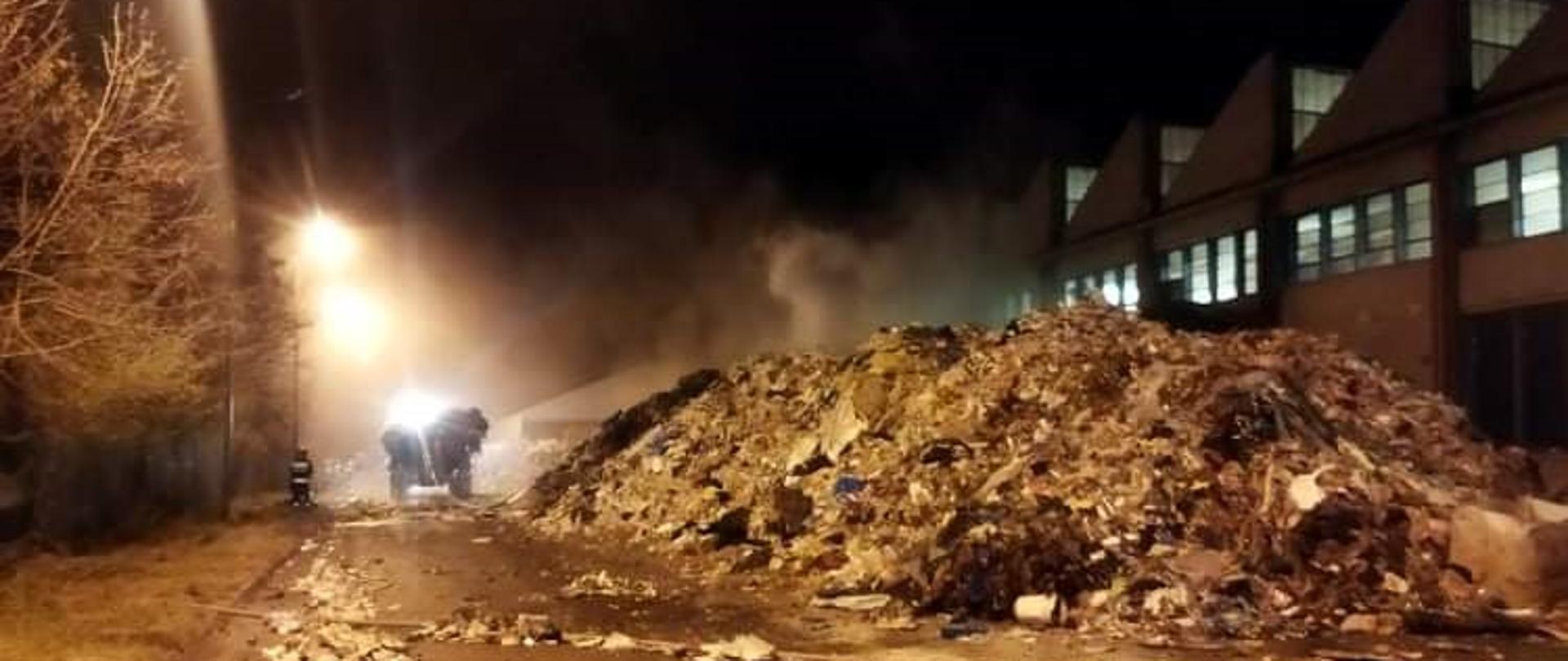 Na zdjęciu z prawej strony widać palącą się hałdę odpadów. Z lewej strony widać ładowarkę typu Fadroma, która przewozi śmieci nieobjęte pożarem w inne miejsce. Koło niej widzimy strażaków, podających prąd wody na palącą się hałdę z odpadami. 