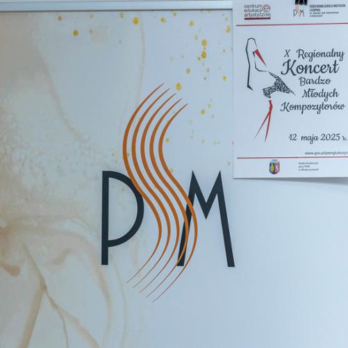 Baner szkolny stojący na scenie, na banerze logo szkoły i przyczepiony w jego górnym prawym rogu plakatem informującym o dziesiątym koncercie bardzo młodych kompozytorów.