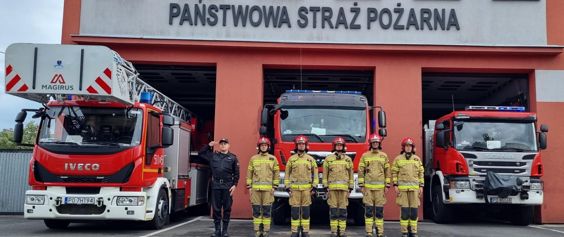 6 strażaków stojących na tle samochodów pożarniczych.