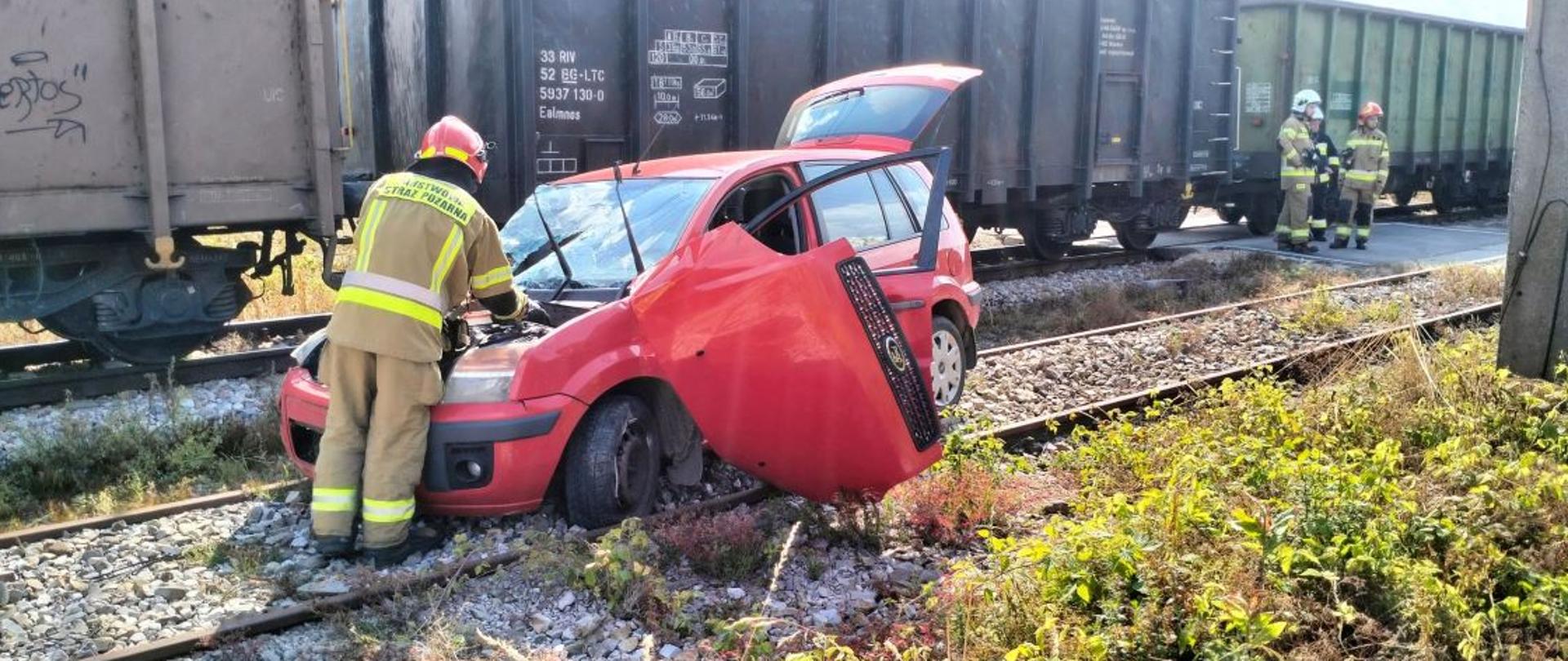 Zdjęcie przedstawia rozbity samochód na torowisku i wagony towarowe pociągu.