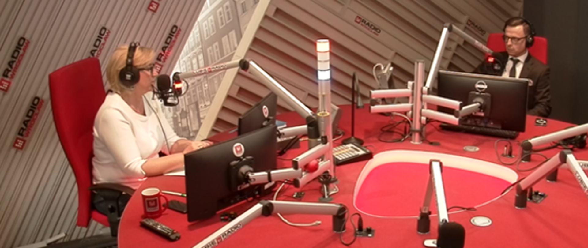 Studio nagraniowe. W centralnej części duży, czerwony stół, na stole ekrany i srebrne ramiona mikrofonowe. 