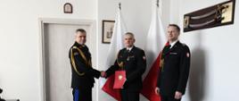Pomorski komendant wojewódzki Państwowej Straży Pożarnej ściska dłoń strażaka trzymającego czerwoną teczkę obok stoi inny funkcjonariusz za nimi stoją flagi Polski. 