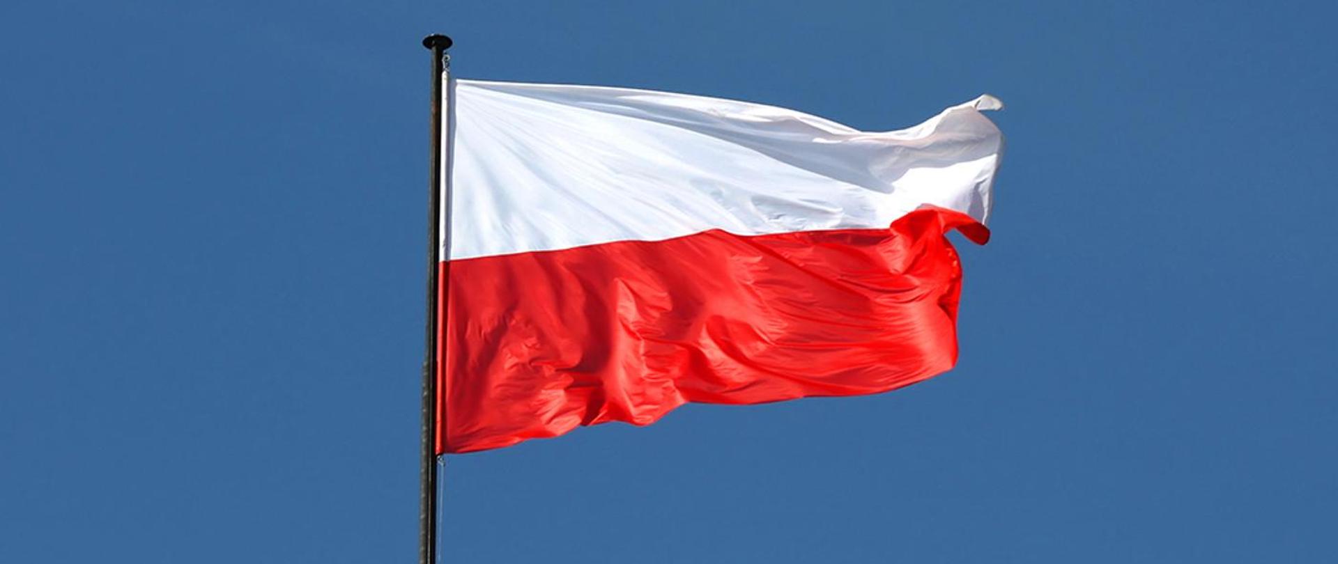 Na zdjęciu widoczna flaga Rzeczypospolitej Polskiej na ciemnym maszcie powiewająca w prawą stronę na tle bezchmurnego nieba.