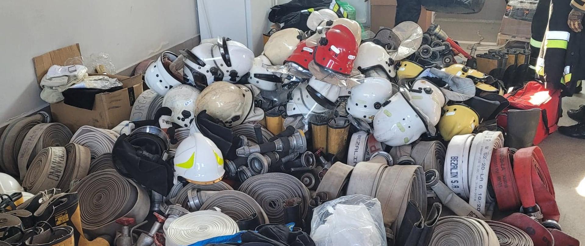 Zdjęcie przedstawia sprzęt strażacki: węże, buty, hełmy, urania znajdujące się na posadzce w garażu, które będą przekazane strażakom z Ukrainy, 
