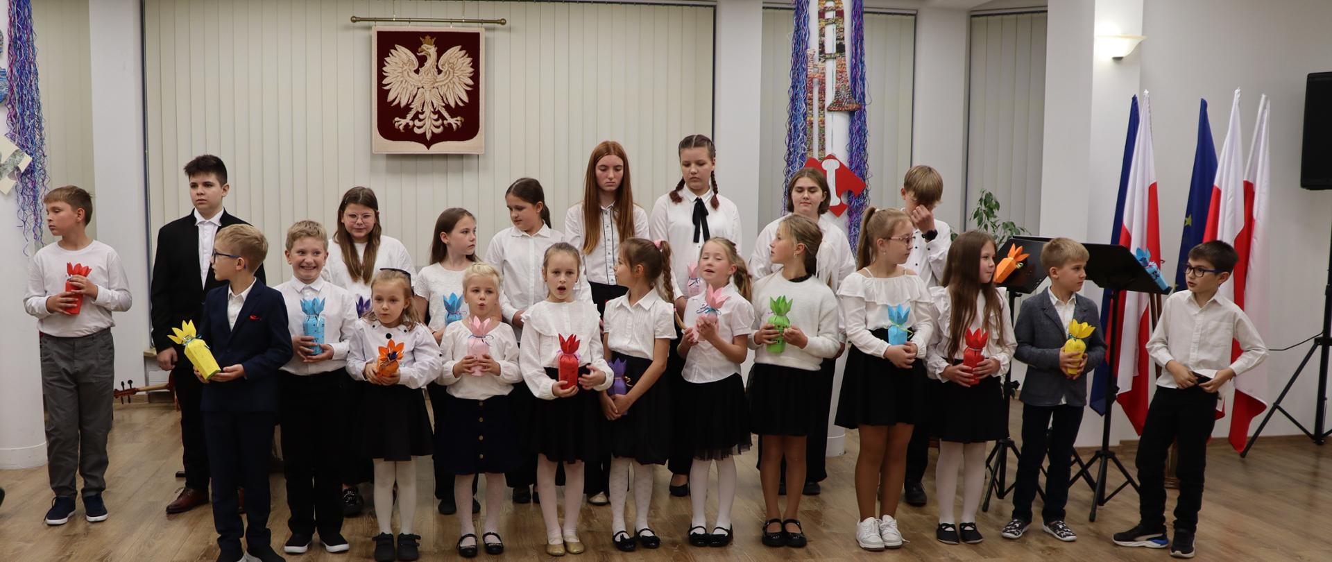 W sali stoją chłopcy i dziewczęta. Dzieci ubrane są w białe koszule i czarne spodnie lub spódnice. W rękach trzymają kolorowe upominki. Za nimi na ścianie wisi godło Polski.