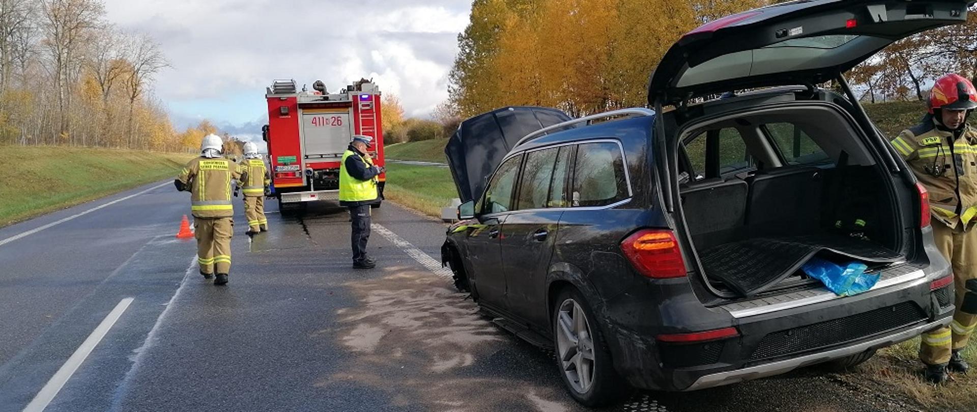 Uszkodzony samochód na poboczu drogi. obok niego strażacy i policjant. W oddali samochód strażacki